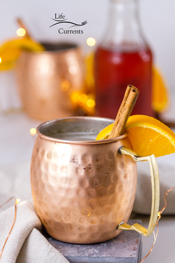 Orange Cinnamon Moscow Mule + Giveaway - This spicy drink can be made non-alcoholic or alcoholic Copper mugs with orange and cinnamon garnishes with a bottle of cinnamon syrup in the background and twinkle lights around