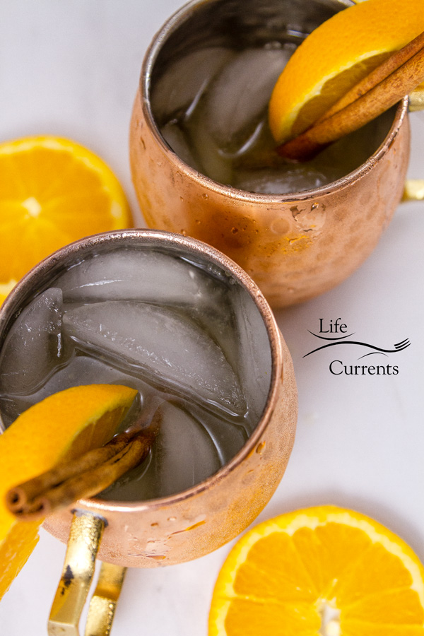 Orange Cinnamon Moscow Mule is a lovely cocktail made with the fall spice flavors of orange and cinnamon. - Orange Cinnamon Moscow Mule + Giveaway top down or flatlay of the Moscow Mule copper cups filled with ice and beverages with orange slices on a white background