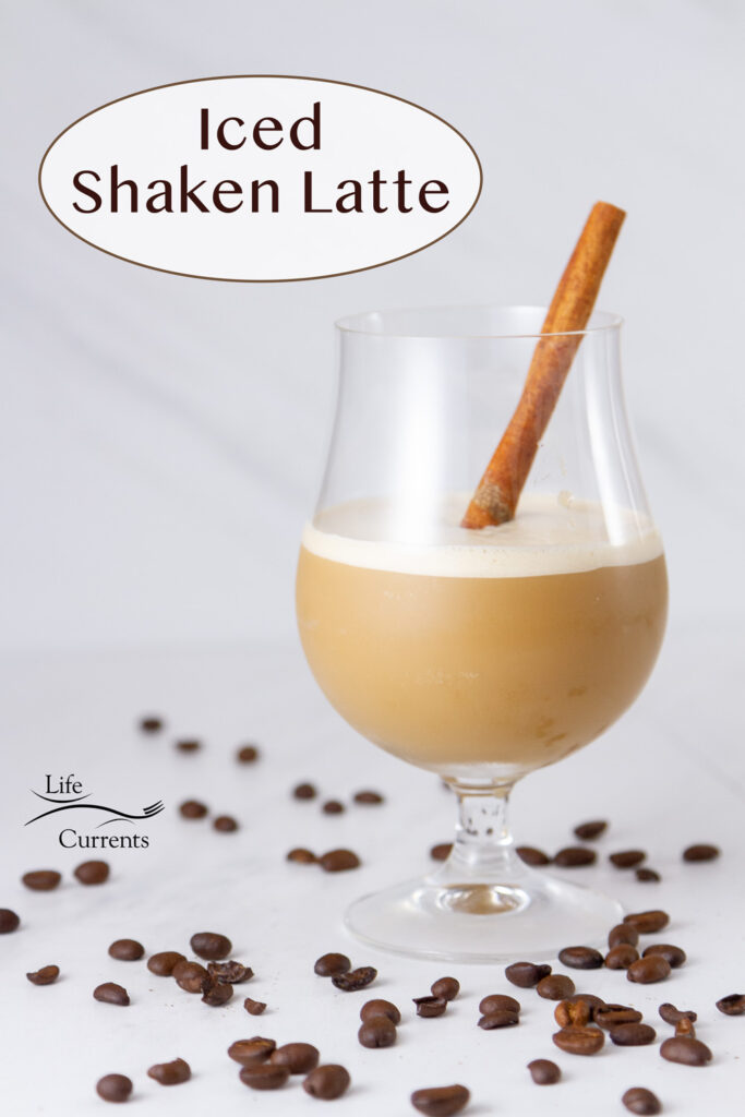 An Iced Shaken Latte in a glass with a cinnamon stick, coffee beans around it.