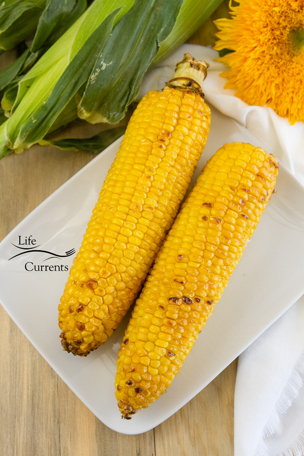 two ears of corn on a white plate with a sunflower and corn still in the husks 