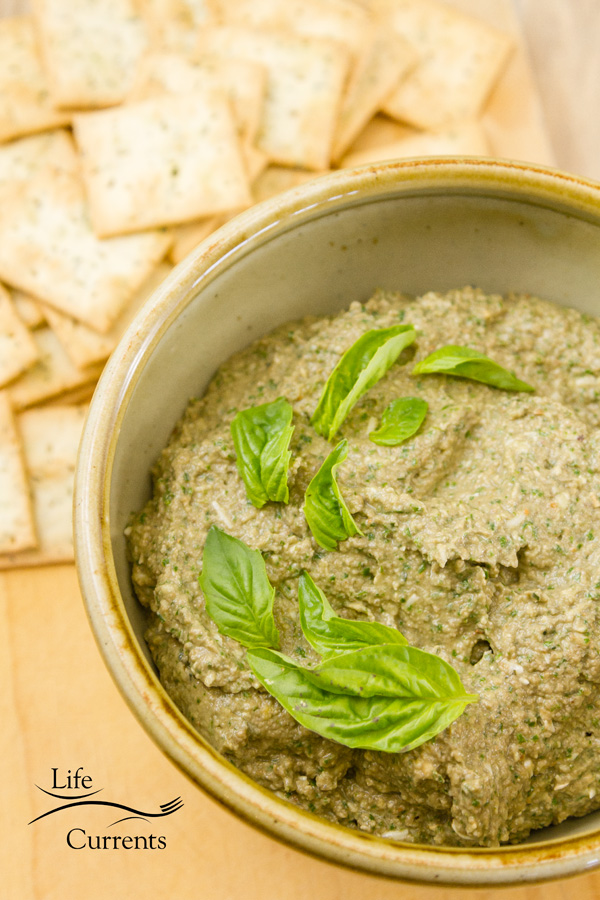 Mushrrom walnut basil pesto spread in an ivory bowl with crackers and fresh green basil leaves on a wood background