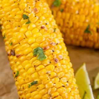 close up on the roasted air fried corn on the cob with green herbs for garnish and lime wedges and another cob in the background