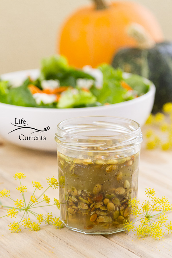 a jar of Toasted Pumpkin Seed Vinaigrette with a large white bowl full of green salad in the back. There are two pumpkins in the very back