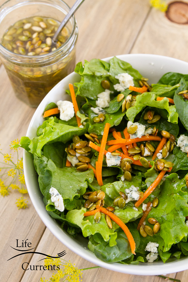 a green salad with lettuce, carrots, blue cheese crumbles, and Toasted Pumpkin Seed Vinaigrette with a jar of Toasted Pumpkin Seed Vinaigrette in the upper left and a spoon in the jar