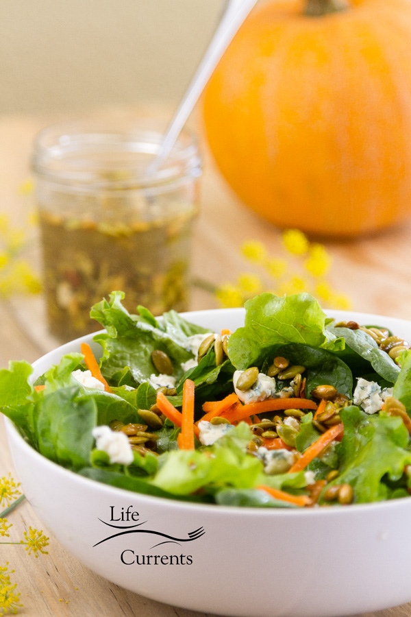 a green salad with a jar of Toasted Pumpkin Seed Vinaigrette and an orange pumpkin in the back