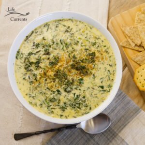 looking down on a big white bowl filled with spinach and artichoke soup and a spoon next to it with some crackers