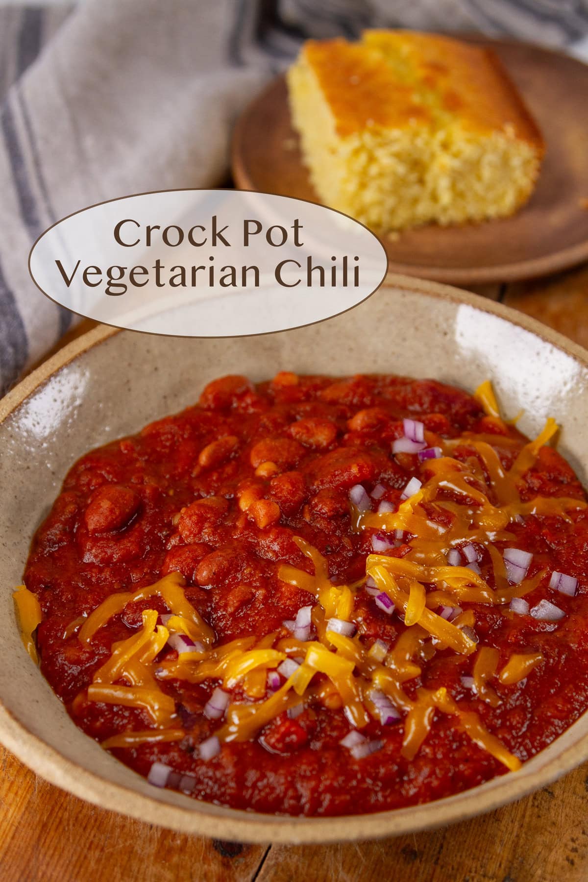 a bowl of chili topped with cheddar and red onions, cornbread in the background.