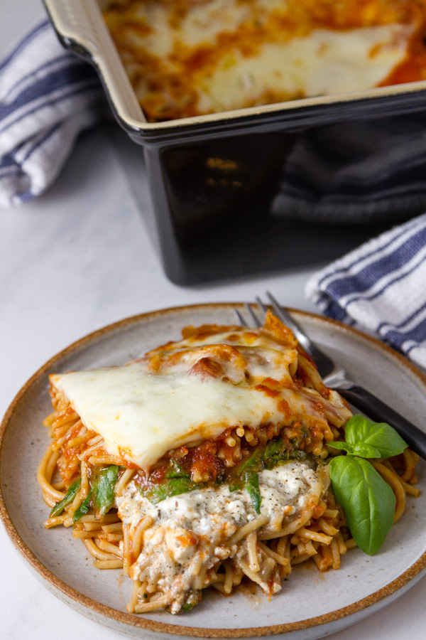 A serving of Spinach Provolone Baked Pasta casserole on a white plate with a garnish of basil and the casserole is in the back with a blue towel