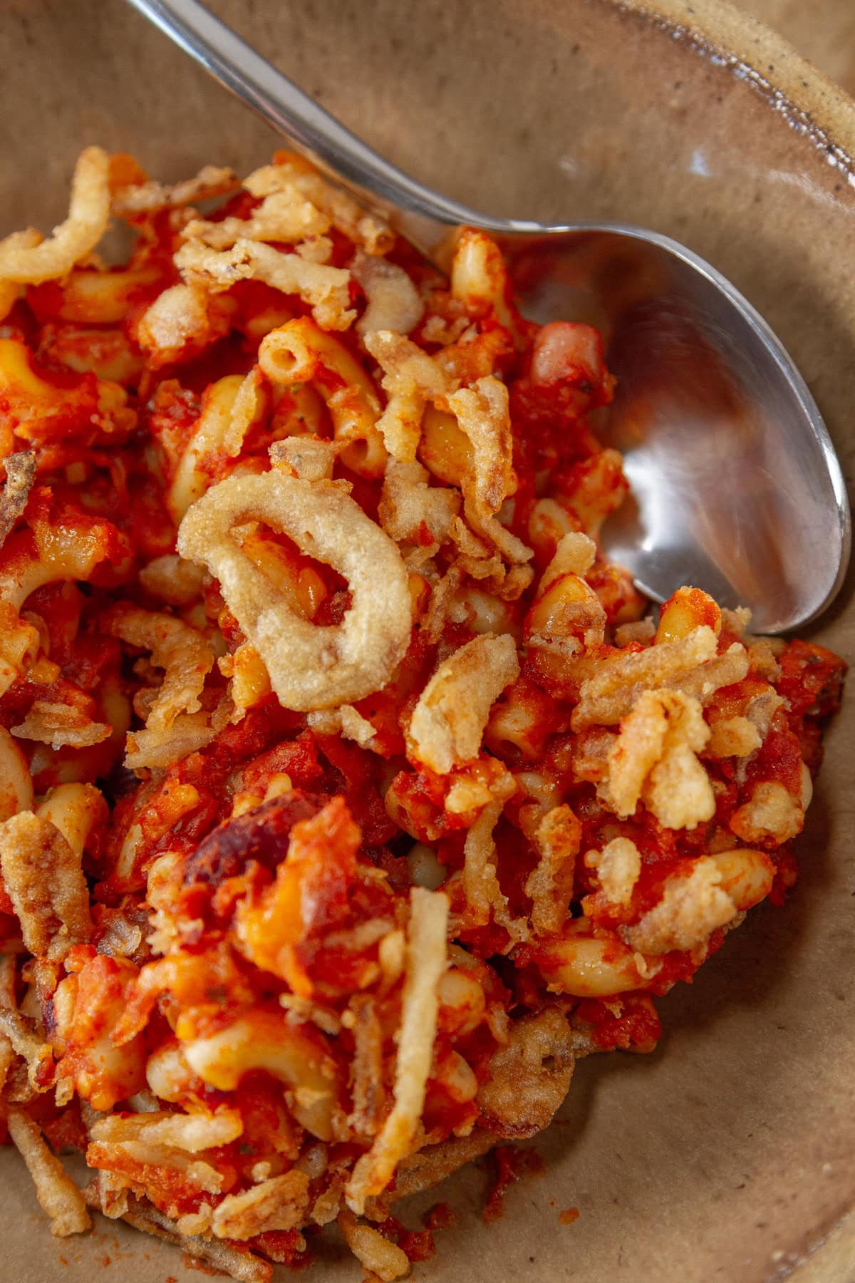 a serving of chili mac in a brown bowl with a spoon in the upper right.