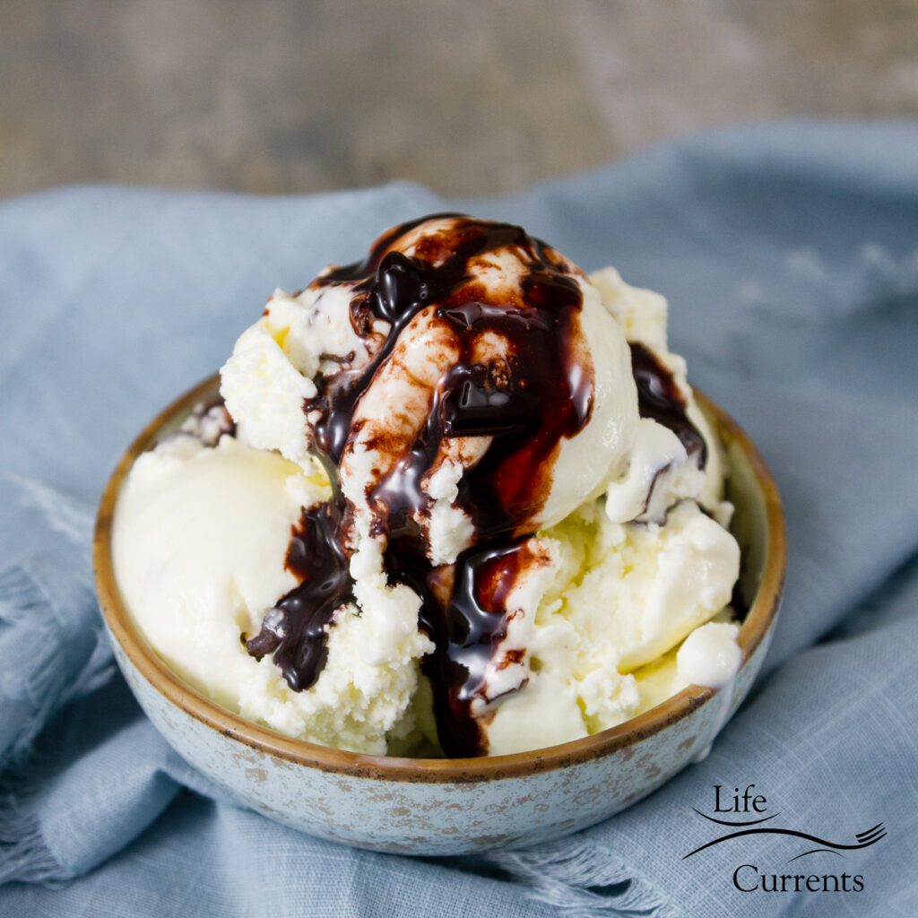 a small bowl of ice cream with chocolate sauce over the top on a blue cloth and the Life Currents logo in the bottom right corner