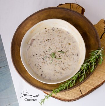 a bowl full of Copycat Souplantation Vegetarian Cream of Mushroom Soup top down shot on a brown plate with a sprig of rosemary in the lower right, square crop