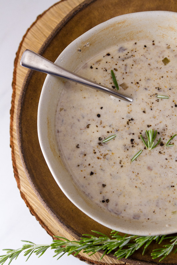 Copycat Souplantation Vegetarian Cream of Mushroom Soup in a bowl with a spoon on a brown plate with a sprig of roasemary at the bottom
