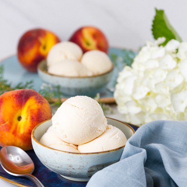 square crop of two bowls of Peach ice cream with peaches and flowers aoround them