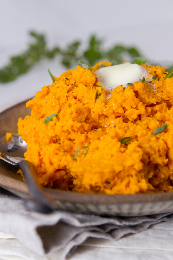 sweet potatoes on a plate with as poon topped with a pat of butter and green onions. Fresh herbs in the background