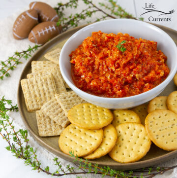 square crop of a bowl of red pepper dip with a plate full of crackers, and some fresh herbs, footballs in the background.