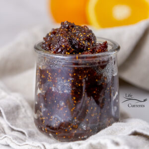 square crop of a glass jar filled with fig jam and some cut oranges in the background.