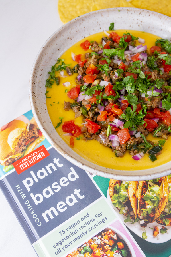 A bowl of appetizer loaded cheese dip with the America&rsquo;s Test Kitchen cookbook &ldquo;Cooking with Plant-Based Meat&rdquo; .