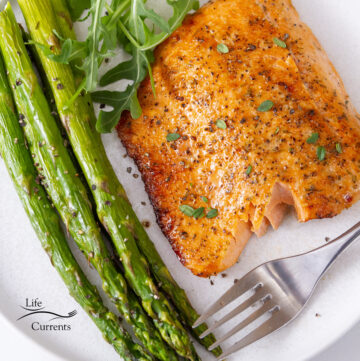 square crop of a cooked piece of salmon on a white plate with asparagus and arugula next to a fork.