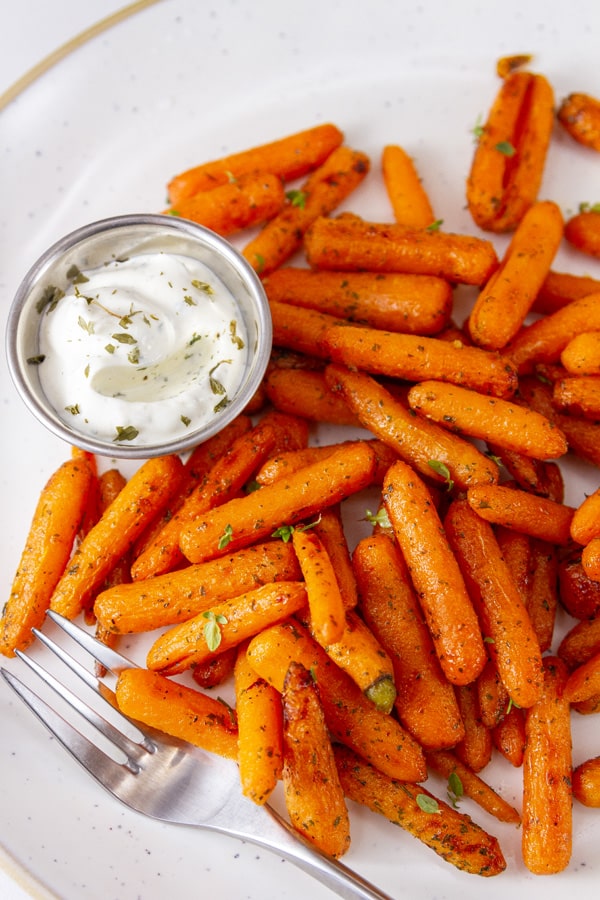 a big batch of cooked carrots on a white plate with a fork and a side dish or ranch dressing.