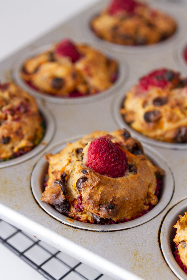muffins in a muffin tray on a cooling rack, each muffin has a raspberry on top.