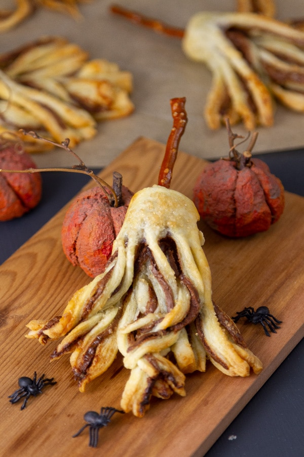 a cookie shaped like a witch's broom leaning against a pumpkin on a wooden serving platter with plastic spiders.