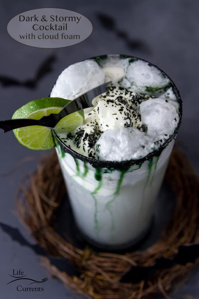 looking down into a Dark and Stormy Cocktail with creamy clouds on top, garnished with a lime wheel and a bat, dry ice floats by in the background.