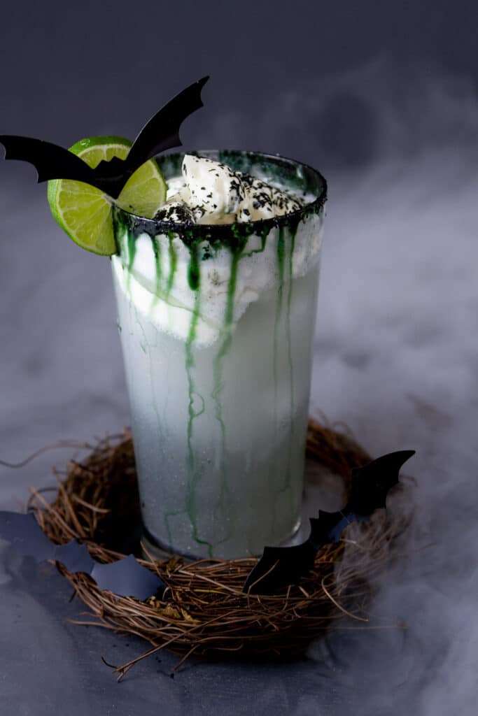 a halloween cocktail with creamy topping, lime wheel, bats, and stick wreath with dry ice smoke in the background.