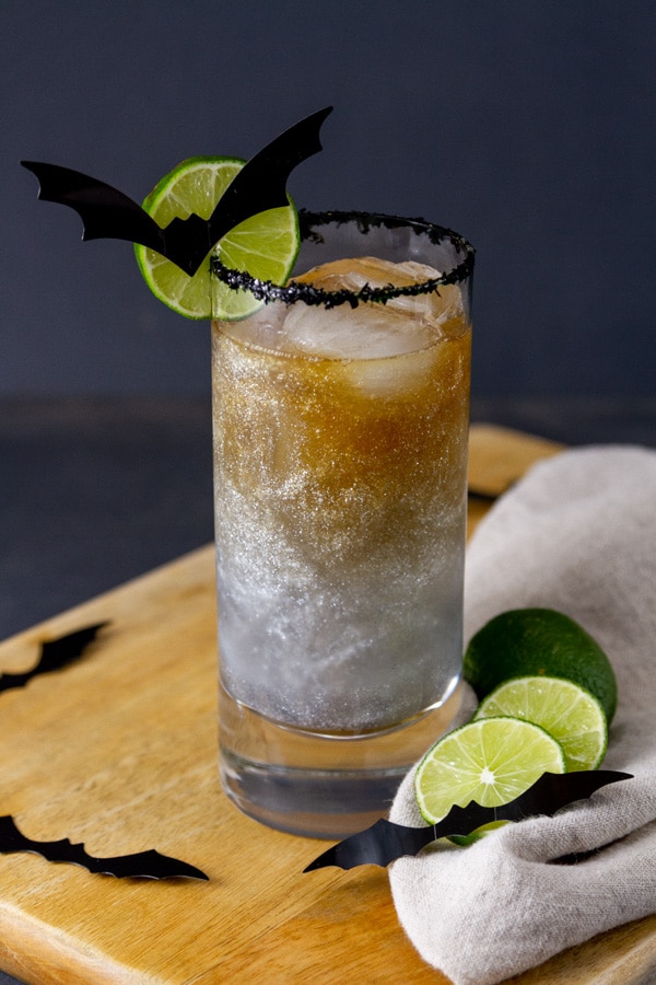 a cocktail drink layered with rum on top, drink sparkles inside, a lime wheel with a bat on the left side, all on a wooden board.