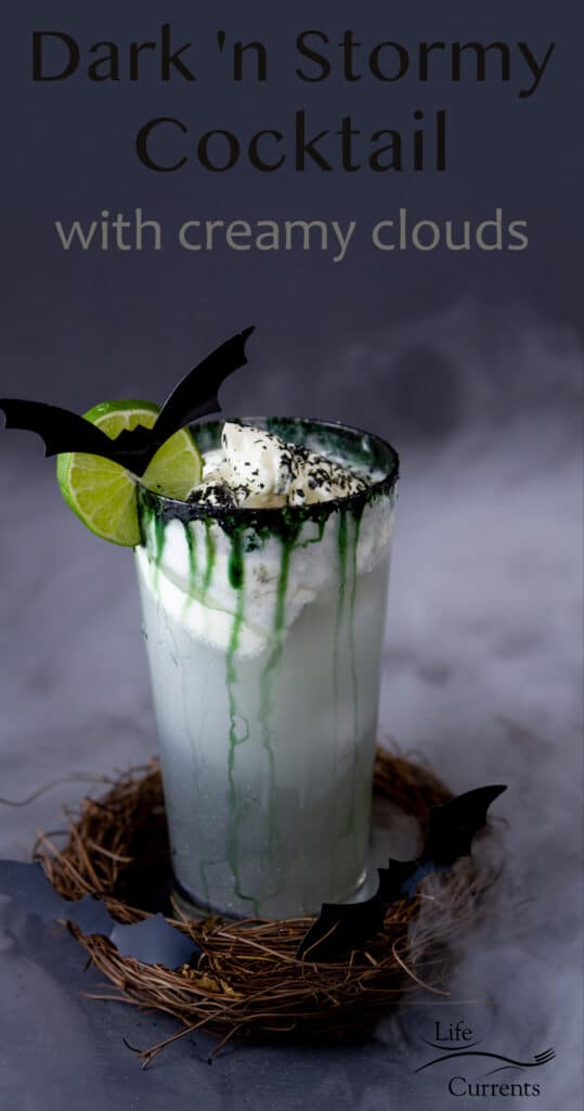 a drink in a glass with whipped cream on top, a lime wheel and a cut out bat, dry ice smoke floats by, title on top: Dark and Stormy Cocktail.