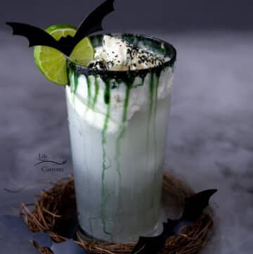 a Dark and Stormy Cocktail drink topped with whipped cream, a black sugar rim, and a lime wheel, cut out bats on the whell and around the base of the drink which is in a stick wreath.