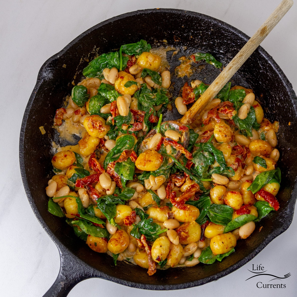 looking fown in a cast iron skillet filled with White Bean Sun-Dried Tomato Gnocchi and a wooden spoon sticking out.