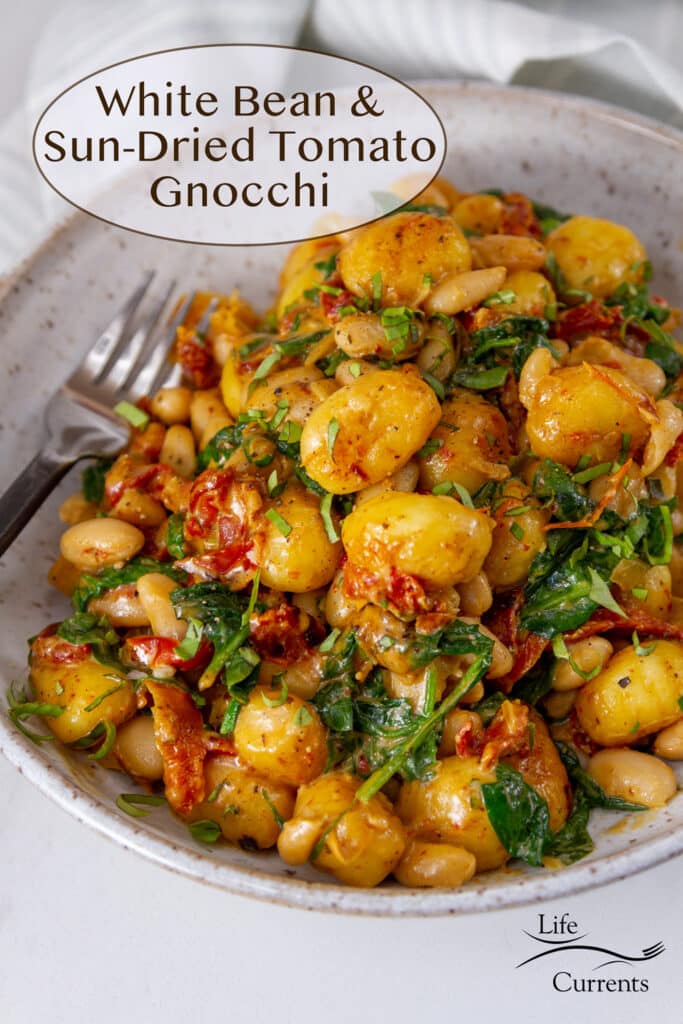 a serving bowl filled with gnocchi, white beans, sun dried tomatoes, and spinach, a fork next to the food, title on top: White Bean Sun-Dried Tomato Gnocchi.