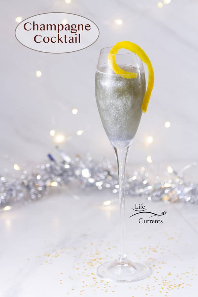 A sparkling Champagne Cocktail in a Champagne glass with a lemon twist in front of silver sparles and twinkle lights.