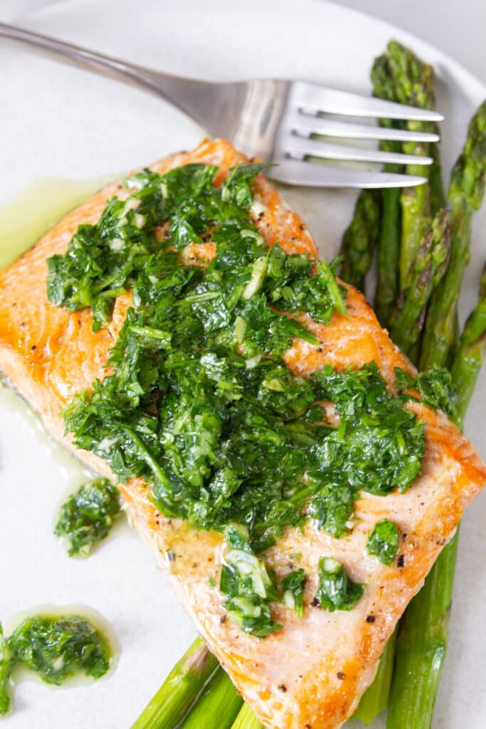 a piece of cooked salmon with chimichurri on top served on top of cooked asparagus with a fork next to it.