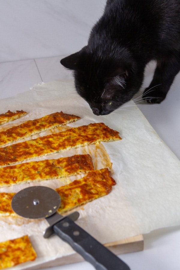 A black cat looking at a plate of cheese sticks with a pizza cutter next to them.