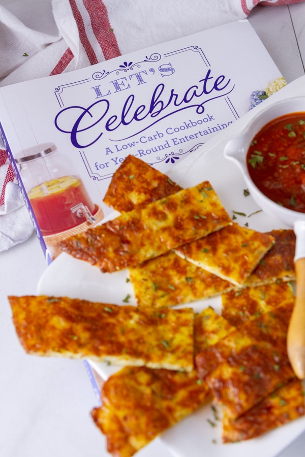 The cookbook Let's Celebrate: A Low-Carb Cookbook for Year-Round Entertaining by Natasha Newton with a plate of cheese sticks on it.