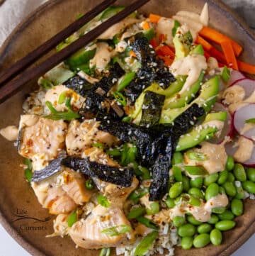 looking down into a sushi bowl with salmon, edamame, seaweed, vegetables, and sesame seeds, chopsticks on the left side.