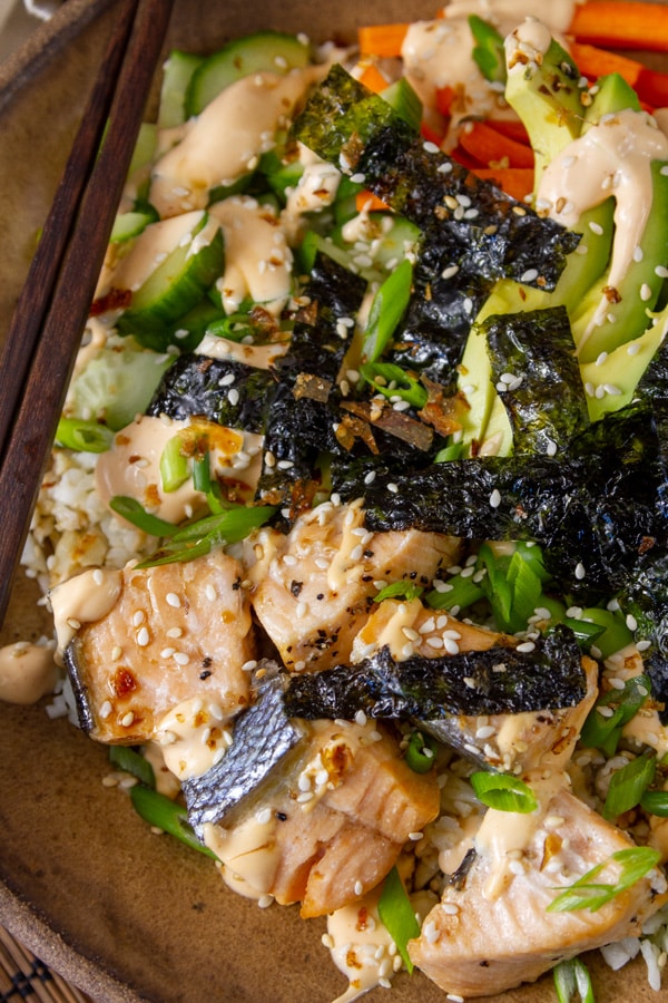 Sushi bowl with salmon, avocado, vegetables, nori, sesame seeds, with chopsticks on the side.