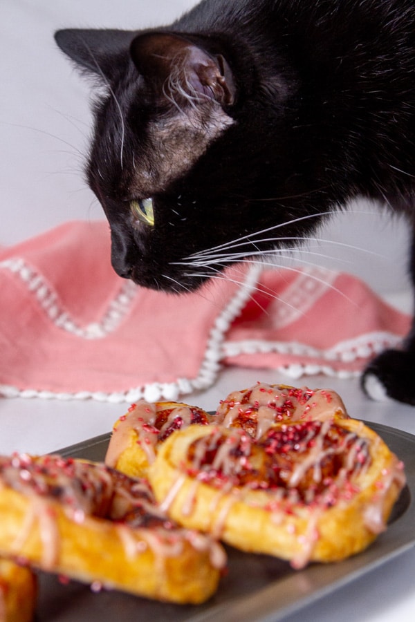a black cat looking at cookies on a pewter plate with a pink napkin in the background.