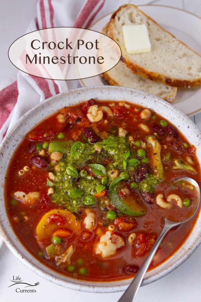 a white bowl and a spoon filled with minestrone soup topped with pesto and basil leaves, a plate with bread slices next to it.