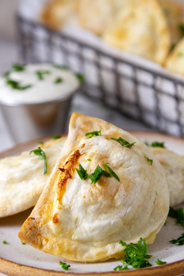 a pierogie cooked in an air fryer on a plate next to ranch dressing in front of a wire basket with more pierogies. 