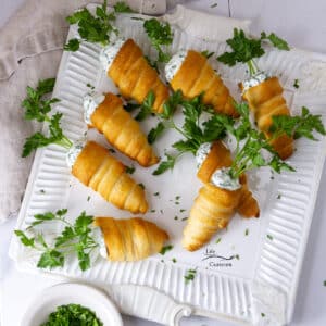 square crop of a bunch of crescent roll carrots stuffed with herb cream cheese on a white serving platter with fresh parsley around.