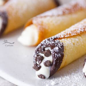 close up on the ends of cannoli cookies, the main focus is a chocolate dipped end sprinkled with chocolate chips, all are dusted with powdered sugar.