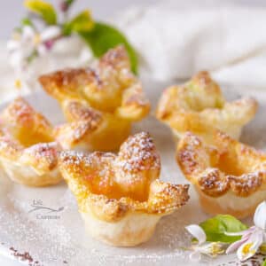 5 puff pastry tarts sprinkled with powdered sugar on a white plate with some lemon tree leaves and flowers around.