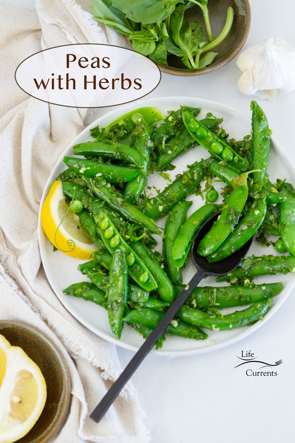 looking down on a plate of sugar snap peas with a lemon wedge and a spoon, a dish of basil and a dish of lemon wedges are next to the plate.