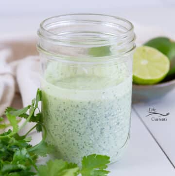 square crop of creamy dressing in a glass jar with fresh cilantro and limes around it.