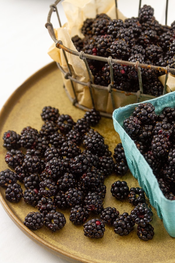 A wire basket and a turquoise paper basket on a brown plate all with blackberries in them.
