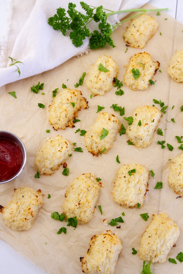 Lots of cauliflower tots on brown parchment paper next to a cup of dipping sauce all garnished with parsley.