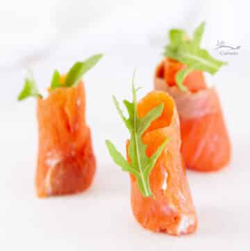 square crop of three smoked salmon rolls with arugula on top on a white plate.