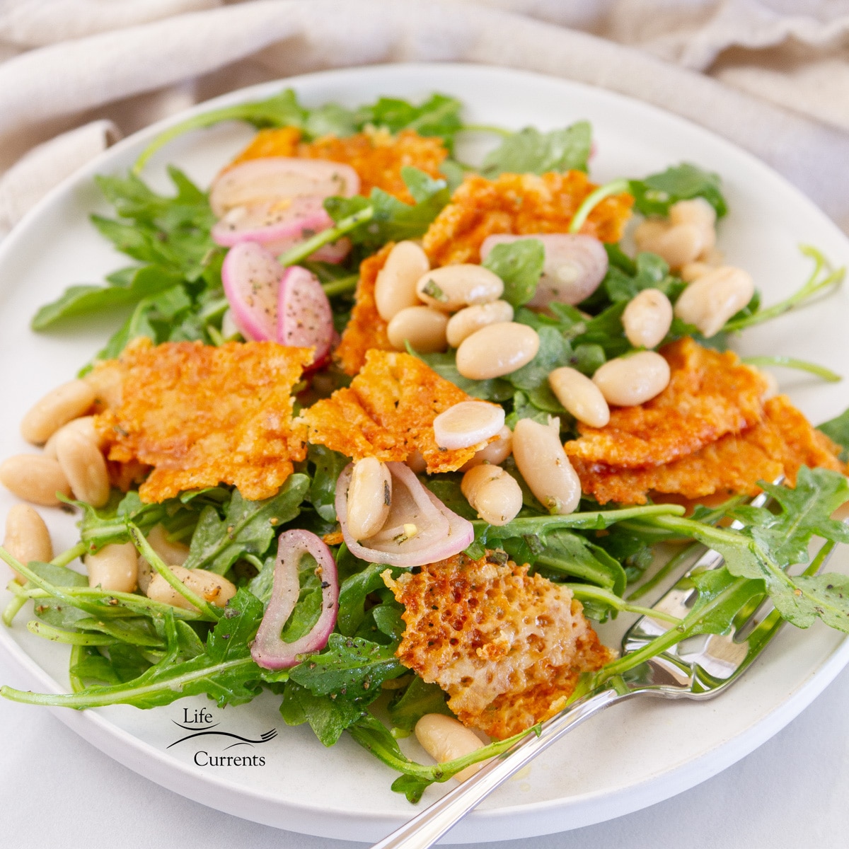 square crop of salad made with arugula white beans and frico (fried cheese) on a white plate with a fork.